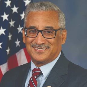 Portrait of a man in a suit and tie in front of a U.S. flag.